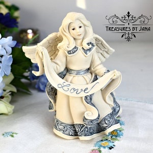 Puede incluir: Una figura de ángel color crema que sostiene una pancarta con la palabra "Love" en escritura cursiva. El ángel tiene alas detalladas y está adornado con detalles azules. La figura es un objeto decorativo.