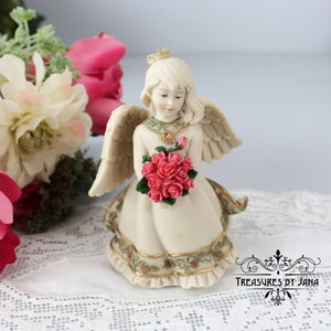 Puede incluir: Una figura de ángel color crema que sostiene un ramo de rosas rosas. El ángel tiene alas detalladas y un vestido decorativo con un estampado floral. La figura tiene un collar dorado y un pequeño lazo en la cabeza. El texto "TREASURES BY JANA" es visible.