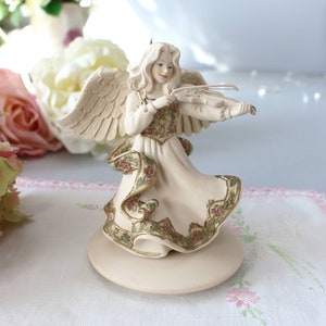 Puede incluir: Una figurita de porcelana blanca de un ángel tocando el violín. El ángel lleva un vestido blanco con detalles florales dorados y verdes. La figurita está sobre una base blanca.