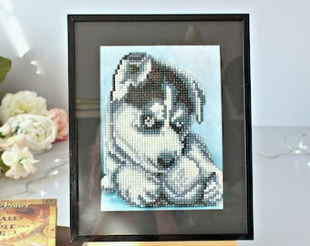 May include: Framed artwork of a puppy, rendered in a mosaic style with black, white, and gray beads. The puppy is set against a light blue background within a black frame. The artwork is displayed on a small wooden easel.