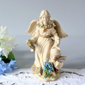 Puede incluir: Una figurita de ángel de porcelana con alas, un vestido blanco fluido y una regadera. El ángel sostiene un rollo y está de pie sobre una base con flores azules y hojas verdes.