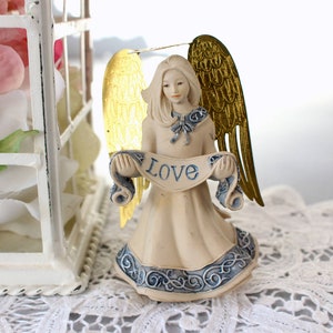 Puede incluir: Una figurita de ángel de porcelana blanca con alas doradas que sostiene un rollo que dice "Love". El ángel lleva un vestido azul y blanco con un estampado floral.