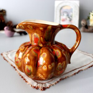 Peut inclure: Pichet décoratif en céramique en forme de citrouille. Le pichet est verni dans des tons de marron, orange et rouge, avec un intérieur blanc et une anse incurvée. Il repose sur un napperon blanc avec une bordure en dentelle.