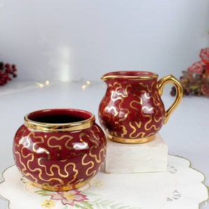 May include: A red and gold ceramic sugar bowl and creamer set with a swirling pattern. The set is on a white doily.