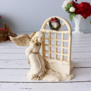 Puede incluir: Una figura de ángel color crema con alas extendidas, arrodillada junto a una ventana enrejada. La ventana está adornada con una pequeña corona y un lazo rojo. La base está esculpida para parecer nieve, creando un tema festivo de vacaciones.