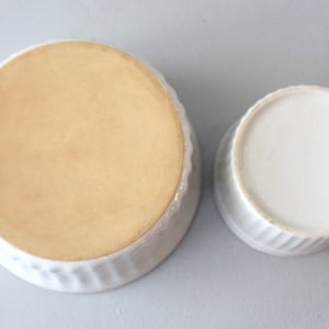 May include: Two white ceramic ramekins, one larger than the other, are shown from a top-down perspective. The larger ramekin has a light tan interior and a ridged exterior. The smaller ramekin has a white interior and a ridged exterior.