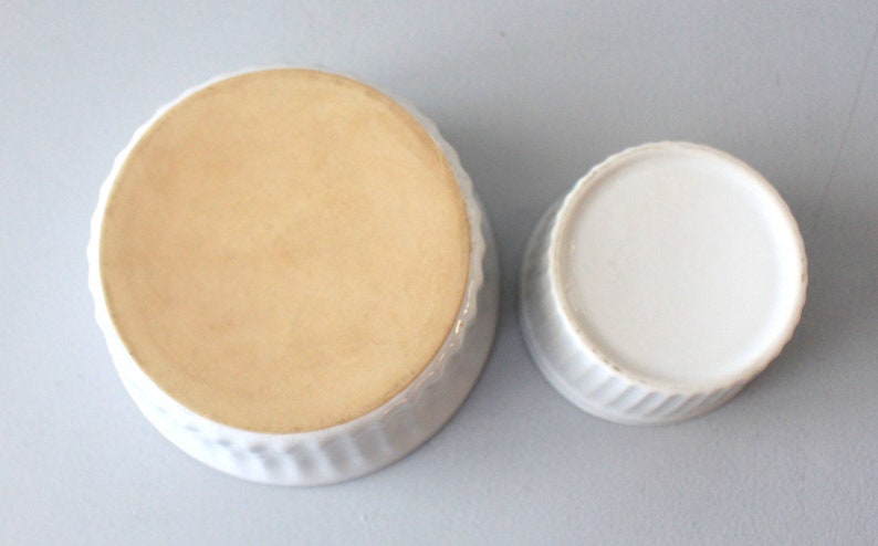 May include: Two white ceramic ramekins, one larger than the other, are shown from a top-down perspective. The larger ramekin has a light tan interior and a ridged exterior. The smaller ramekin has a white interior and a ridged exterior.