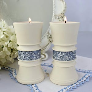 Puede incluir: Dos candelabros de color marfil con velas encendidas. Cada uno tiene una banda floral azul. Los candelabros están sobre una superficie blanca con ribete azul, lo que sugiere un elemento decorativo para el hogar.