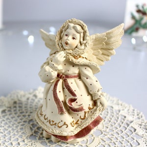 Puede incluir: Una figurita de ángel de porcelana blanca con un vestido de lunares rojos y blancos, una cinta roja y detalles dorados. El ángel tiene alas y lleva una capucha de piel blanca. La figurita está sobre un encaje de ganchillo blanco.