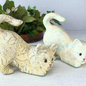 K&ouml;nnte beinhalten: Zwei Keramik-Katzenfiguren. Eine Katze ist cremefarben mit einem grauen Blumenmuster. Die andere Katze ist cremefarben mit hellblauen Akzenten. Beide Katzen befinden sich in einer spielerischen Pose, mit gekr&uuml;mmten Schw&auml;nzen.