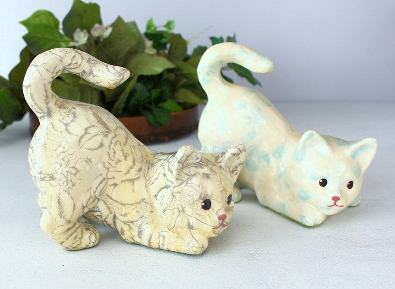 K&ouml;nnte beinhalten: Zwei Keramik-Katzenfiguren. Eine Katze ist cremefarben mit einem grauen Blumenmuster. Die andere Katze ist cremefarben mit hellblauen Akzenten. Beide Katzen befinden sich in einer spielerischen Pose, mit gekr&uuml;mmten Schw&auml;nzen.