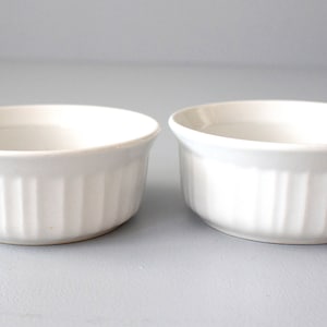May include: Two white ceramic ramekins with vertical ribbed sides. The ramekins have a slightly flared rim and are set against a light grey background. The number "#2" is in the lower left corner.