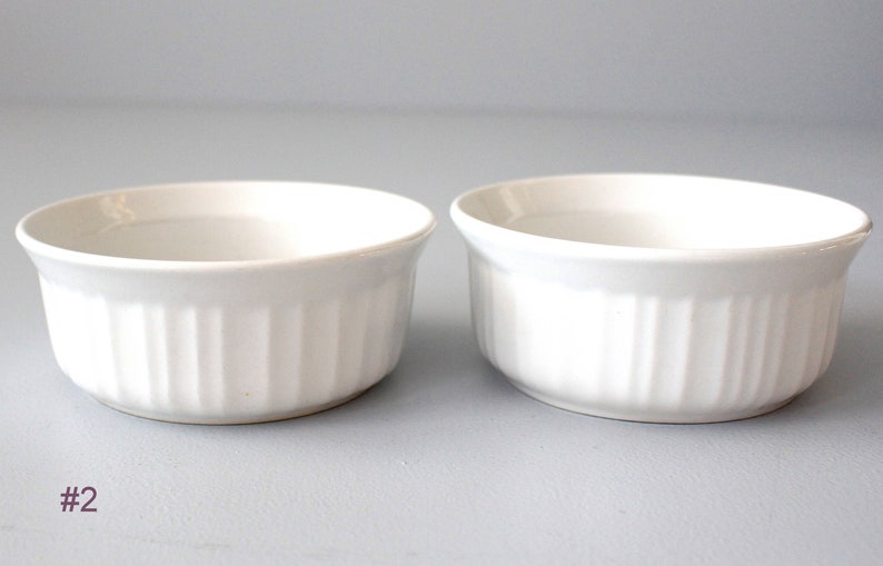 May include: Two white ceramic ramekins with vertical ribbed sides. The ramekins have a slightly flared rim and are set against a light grey background. The number "#2" is in the lower left corner.