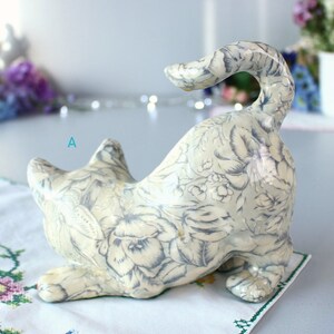 K&ouml;nnte beinhalten: Keramik-Katzenfigur in spielerischer Pose, mit cremefarbenem Grund und zartgrauem Blumenmuster. Die Katze ist gebogen, der Schwanz ist erhoben. Die Figur steht auf einer wei&szlig;en Oberfl&auml;che mit floralem Stickdesign.