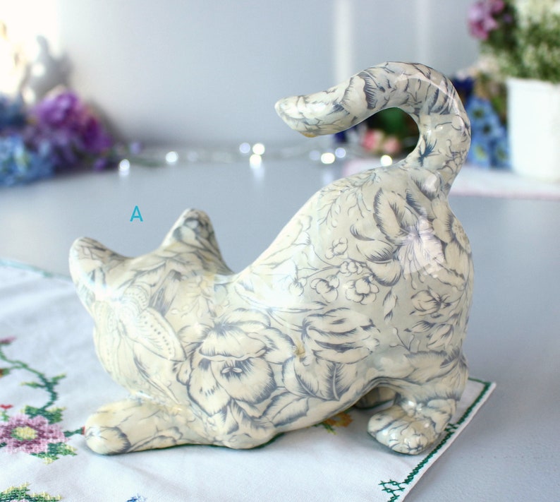 K&ouml;nnte beinhalten: Keramik-Katzenfigur in spielerischer Pose, mit cremefarbenem Grund und zartgrauem Blumenmuster. Die Katze ist gebogen, der Schwanz ist erhoben. Die Figur steht auf einer wei&szlig;en Oberfl&auml;che mit floralem Stickdesign.