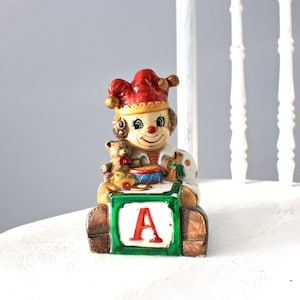 May include: A ceramic clown figurine holding a teddy bear and a drum, sitting on a green block with the letter 'A' on it.