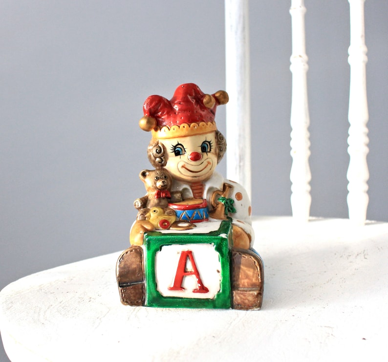 May include: A ceramic clown figurine holding a teddy bear and a drum, sitting on a green block with the letter 'A' on it.