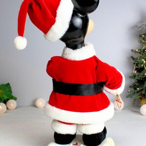 May include: A red and white plush Mickey Mouse dressed as Santa Claus. He is standing on a white base with a red button. The figure is approximately 18 inches tall.