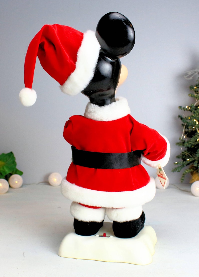 May include: A red and white plush Mickey Mouse dressed as Santa Claus. He is standing on a white base with a red button. The figure is approximately 18 inches tall.