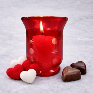 May include: A burning red candle in a decorative glass holder with star and dot designs. Heart-shaped chocolates and plush hearts in red, white, and brown are beside the candle. The candle flame is bright.