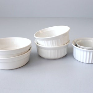 May include: Several white ceramic ramekins of different sizes. Some have a fluted design, others are smooth and rounded. The ramekins are stacked and arranged on a light grey surface. Suitable for baking and serving individual portions, these ramekins are a kitchen essential.