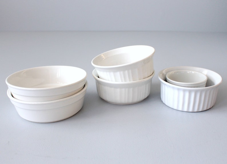 May include: Several white ceramic ramekins of different sizes. Some have a fluted design, others are smooth and rounded. The ramekins are stacked and arranged on a light grey surface. Suitable for baking and serving individual portions, these ramekins are a kitchen essential.