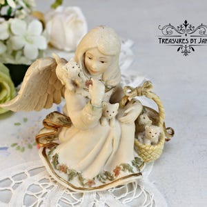 Puede incluir: Una figurita de porcelana de un ángel con alas, sosteniendo una cesta de gatitos. El ángel lleva un vestido blanco y tiene el pelo largo y rubio. La cesta está tejida y tiene un asa marrón. La figurita está sentada sobre un mantel de encaje blanco.