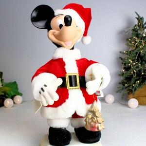 May include: A plush Mickey Mouse dressed as Santa Claus, wearing a red suit with white trim and a black belt. He is holding a gold-colored ornament with a Christmas scene.