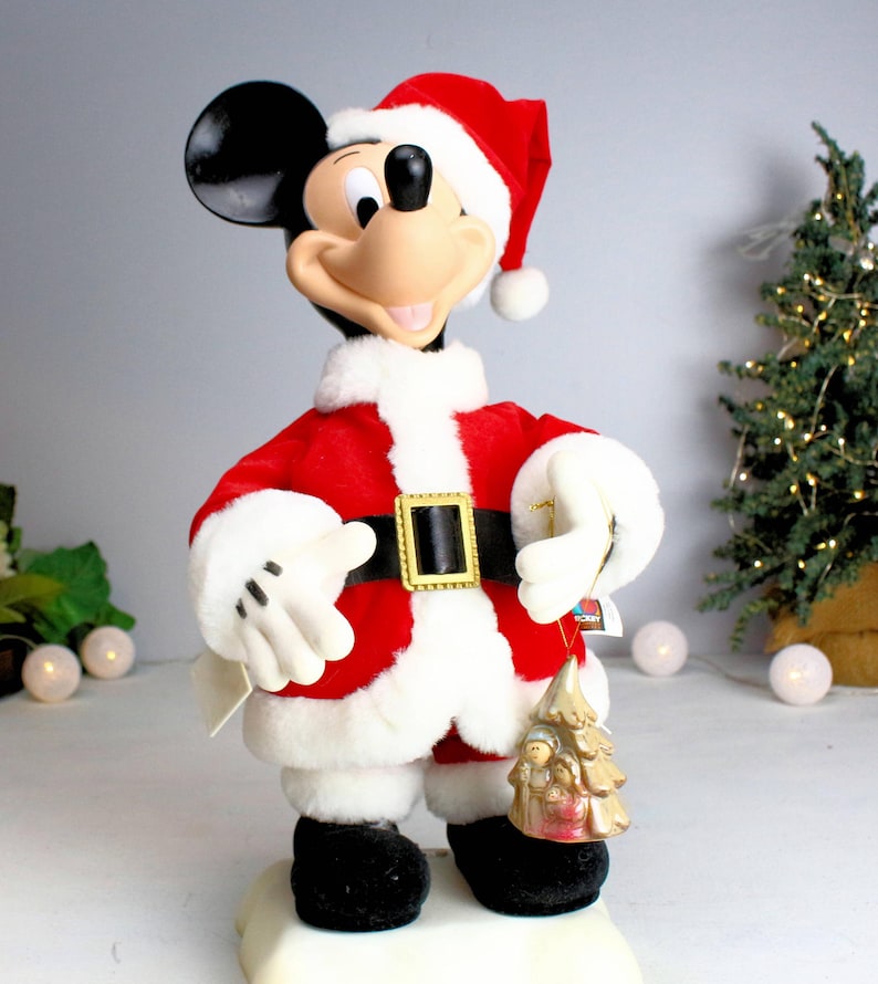 May include: A plush Mickey Mouse dressed as Santa Claus, wearing a red suit with white trim and a black belt. He is holding a gold-colored ornament with a Christmas scene.