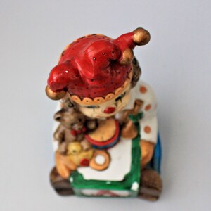 May include: A ceramic clown figurine with a red and white jester hat, holding a teddy bear and a toy drum. The clown is sitting on a colourful block with the letter A.