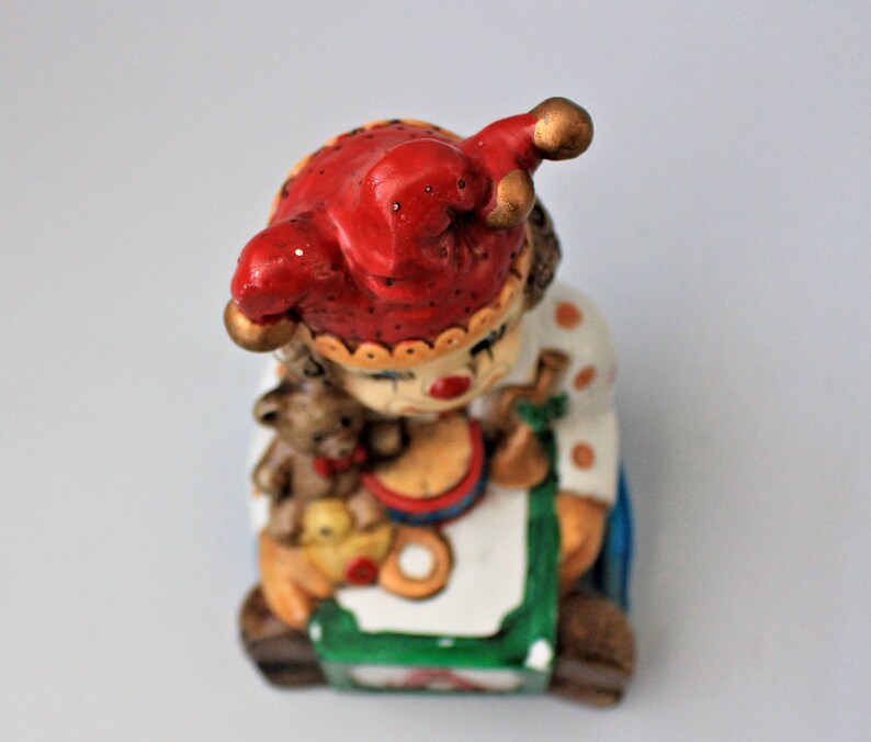 May include: A ceramic clown figurine with a red and white jester hat, holding a teddy bear and a toy drum. The clown is sitting on a colourful block with the letter A.