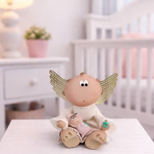 Könnte beinhalten: Handgefertigte Engelsfigur mit goldfarbenen Flügeln, die eine Babypuppe hält. Der Engel trägt ein weißes Gewand und hat einen Schnuller. Die Figur sitzt auf einer weißen Oberfläche, mit einem Kinderbett im Hintergrund.