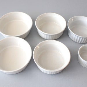 May include: Six white ceramic ramekins of varying sizes are arranged on a light surface. The ramekins have a smooth, glossy finish, with some featuring a ribbed design on the exterior. These small, round dishes are ideal for baking and serving individual portions.
