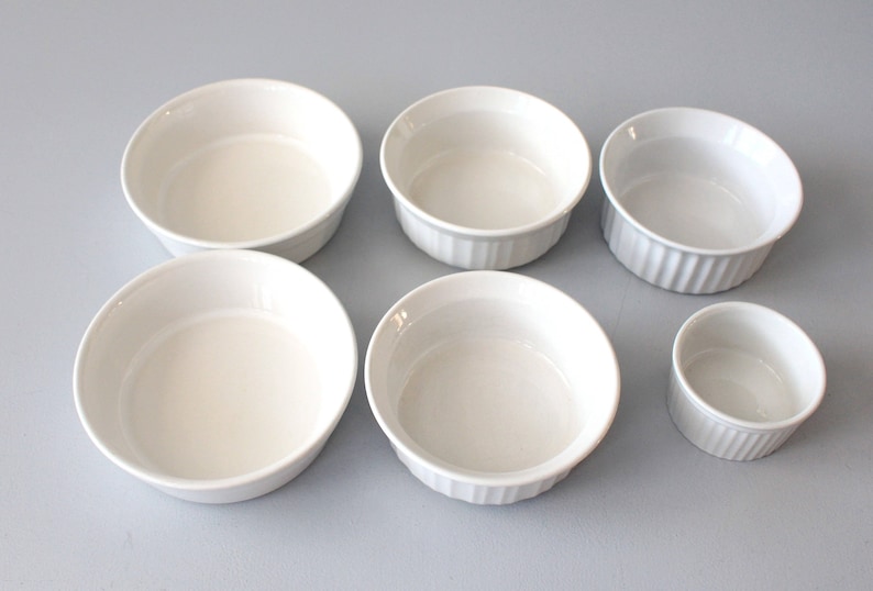May include: Six white ceramic ramekins of varying sizes are arranged on a light surface. The ramekins have a smooth, glossy finish, with some featuring a ribbed design on the exterior. These small, round dishes are ideal for baking and serving individual portions.