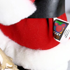 May include: A red and white Santa Claus plush toy with a black belt and a Mickey Mouse tag that reads "Disney, Made in China, Fabriqu&eacute; en Chine".