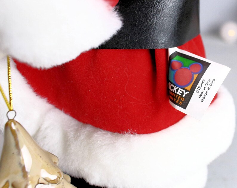 May include: A red and white Santa Claus plush toy with a black belt and a Mickey Mouse tag that reads "Disney, Made in China, Fabriqu&eacute; en Chine".