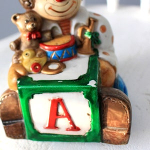 May include: A ceramic clown figurine holding a toy drum, a teddy bear, and a rubber duck. The clown is sitting on a green and white block with the letter 'A' on it.