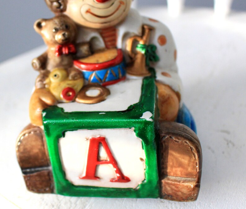 May include: A ceramic clown figurine holding a toy drum, a teddy bear, and a rubber duck. The clown is sitting on a green and white block with the letter 'A' on it.