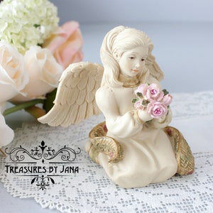 Puede incluir: Una figurita de ángel de porcelana blanca arrodillada con las alas extendidas, sosteniendo un ramo de rosas rosas. El ángel lleva un vestido blanco y tiene el pelo largo y rubio. La figurita está sentada sobre un encaje blanco.