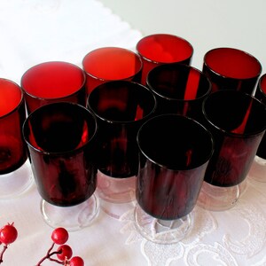 May include: A collection of ten red-tinted glass tumblers with clear, circular bases. The glasses have a slightly angled design and are arranged on a white, textured surface. The colour of the glasses varies from a deep red to a darker, almost black hue.