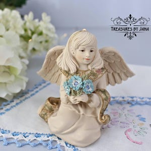 Puede incluir: Figura de ángel color crema arrodillada sosteniendo un ramo de flores azules. El ángel tiene alas detalladas y viste un vestido con detalles dorados y florales. El texto "Treasures by Jana" es visible en la esquina superior derecha.