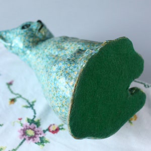 Pode incluir: Uma figura de gato de cer&acirc;mica azul com um fundo de feltro verde. O gato est&aacute; coberto com um padr&atilde;o floral de pequenas flores azuis.