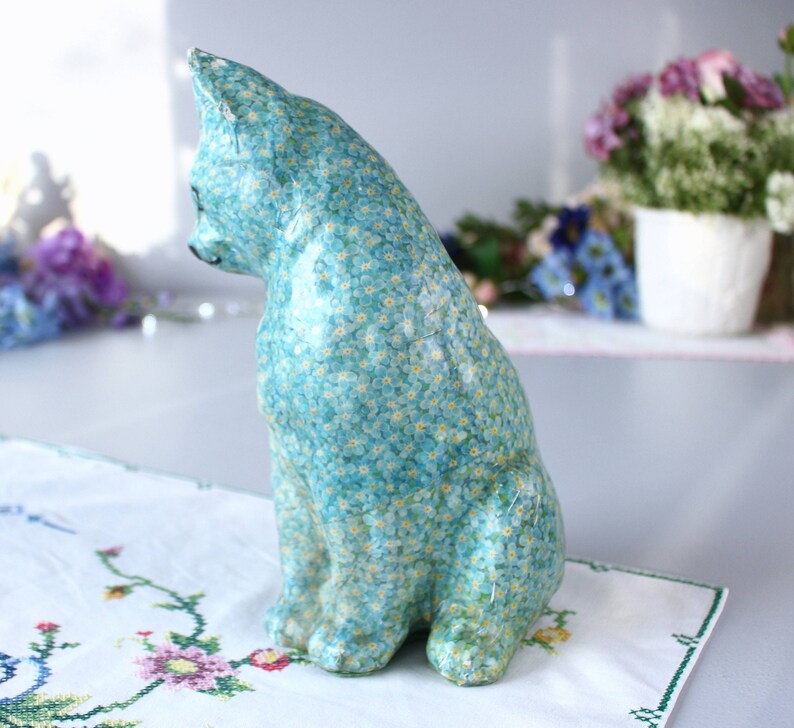 Pode incluir: Uma figura de cer&acirc;mica azul de um gato com um padr&atilde;o floral de miosotis. O gato est&aacute; sentado com a cabe&ccedil;a virada para o lado.