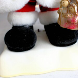 May include: A plush Santa Claus figure wearing a red suit and white trim, standing on a white base. He is holding a ceramic figurine of the nativity scene.