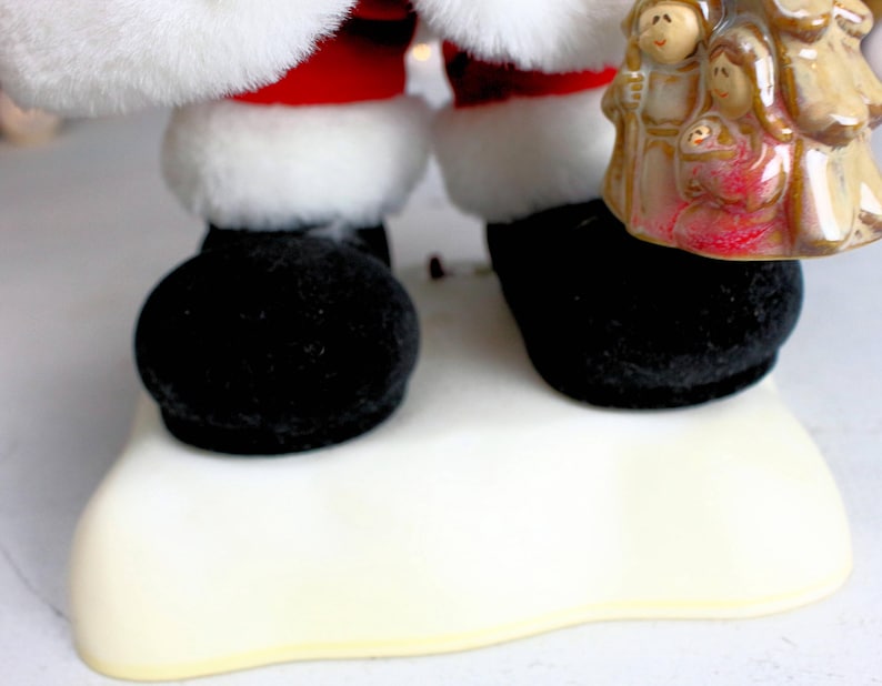 May include: A plush Santa Claus figure wearing a red suit and white trim, standing on a white base. He is holding a ceramic figurine of the nativity scene.