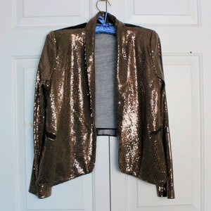 May include: A bronze sequined blazer with a draped front and black trim. The open-front jacket is hanging on a blue hanger against a white door. The blazer has a shimmering, textured appearance.