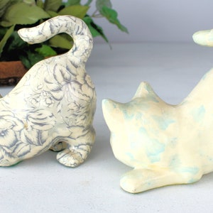 K&ouml;nnte beinhalten: Zwei Keramik-Katzenfiguren in spielerischer Pose. Eine Katze ist cremefarben mit einem grauen Blumenmuster, die andere ist cremefarben mit blauen Akzenten. Beide haben einen gebogenen Schwanz und stehen auf einer wei&szlig;en Oberfl&auml;che.