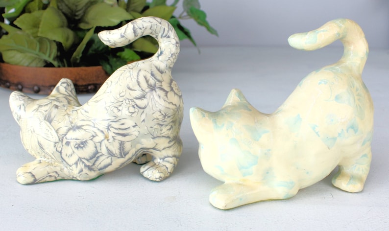 K&ouml;nnte beinhalten: Zwei Keramik-Katzenfiguren in spielerischer Pose. Eine Katze ist cremefarben mit einem grauen Blumenmuster, die andere ist cremefarben mit blauen Akzenten. Beide haben einen gebogenen Schwanz und stehen auf einer wei&szlig;en Oberfl&auml;che.