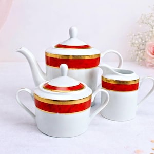 Puede incluir: Un juego de té de porcelana blanca con detalles rojos y dorados. El juego incluye una tetera, un azucarero y una jarra de crema. La tetera tiene un pico curvo y una tapa con un pequeño pomo. El azucarero tiene dos asas y una tapa.