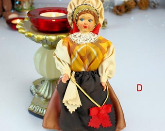 May include: A vintage doll dressed in traditional attire, featuring a yellow and brown patterned bodice, a black skirt, and a cream-colored bonnet. The doll has blue eyes and red accents. A red candle holder is in the background.